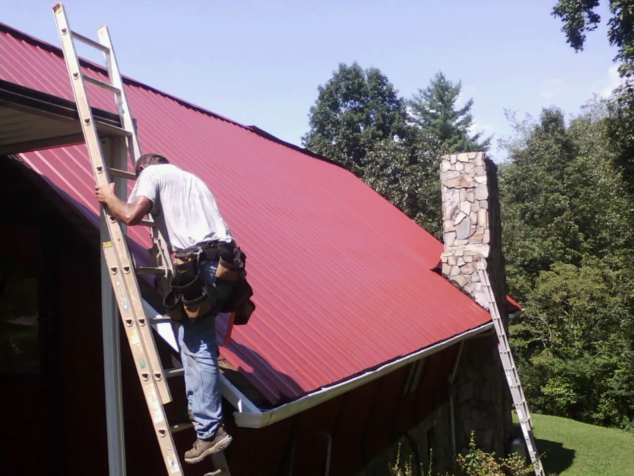 Roofer on a ladder beside a red metal roof and stone chimney — Soffit & Fascia Repair by Roofing Brothers in Davey